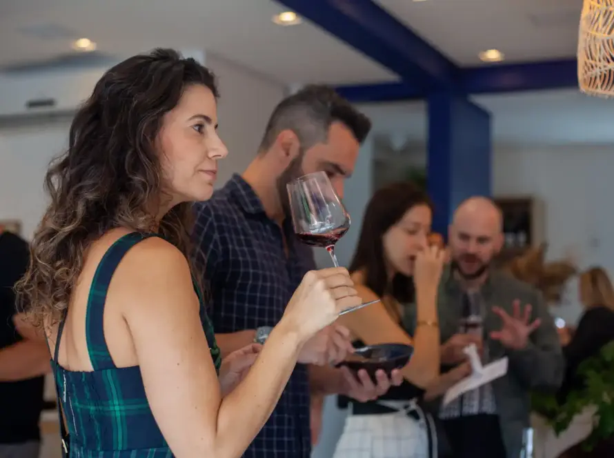 Mulher em evento social segurando taça de vinho tinto, com amigos ao fundo, em ambiente elegante e social.