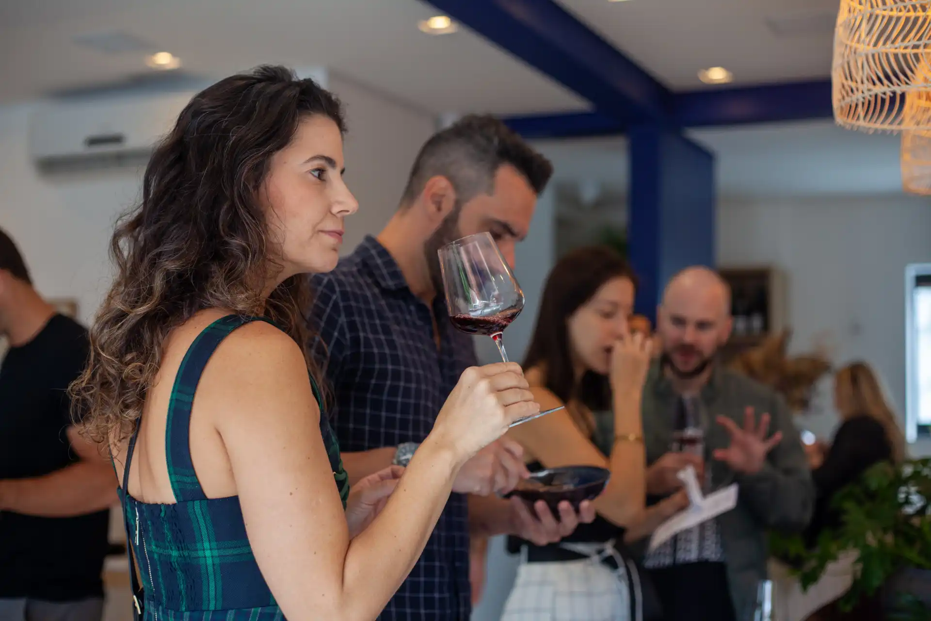 Mulher em evento social segurando taça de vinho tinto, com amigos ao fundo, em ambiente elegante e social.
