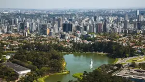 Vista panorâmica de São Paulo com a cidade ao fundo e o Parque Ibirapuera em primeiro plano, incluindo o espelho d'água e o Monumento às Bandeiras, em uma manhã ensolarada, mostrando o contraste entre a natureza e a urbanização da capital paulista.
