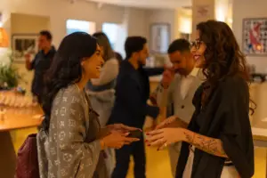Duas mulheres conversando e sorrindo durante uma festa ou evento social no winebar da Cellar Cave, com várias pessoas ao fundo em ambiente bem iluminado.